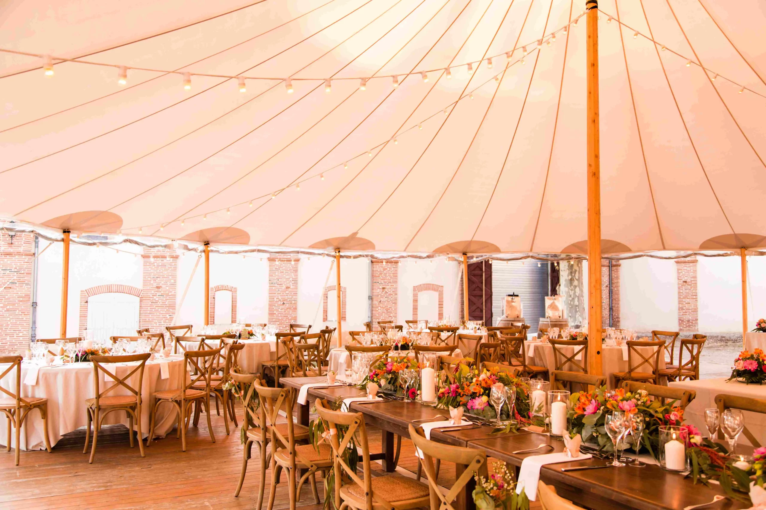 Salle sous tente avec tables en bois décorées de compositions florales pour un mariage élégant – Poppy & Daisy